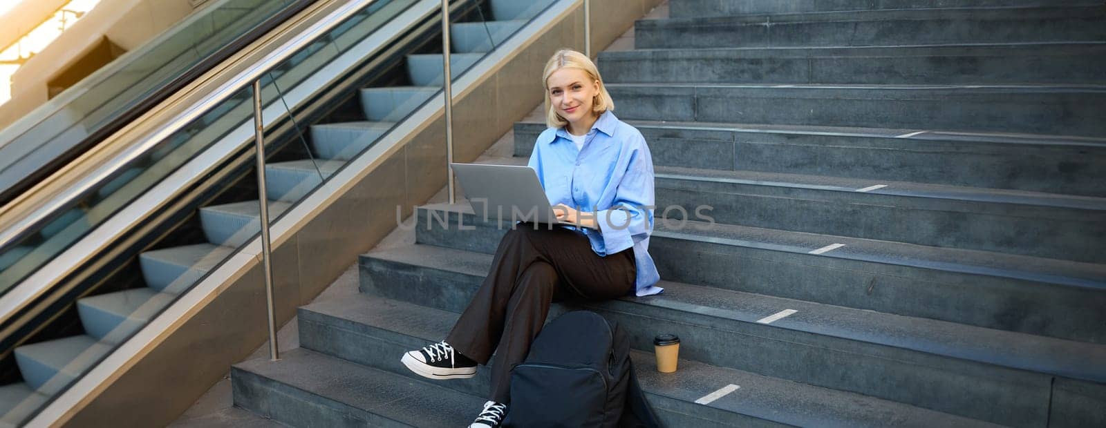 Portrait of young woman, student sitting outdoors on stairs with coffee, using laptop, working on project, looking at camera and smiling.