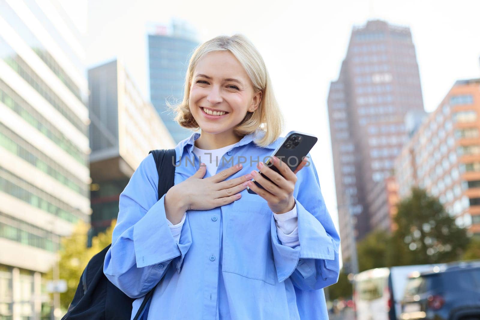 Close up portrait of stylish young blonde woman, standing on street, checking her mobile phone, using smartphone app to get around town, looking at online map for directions by Benzoix
