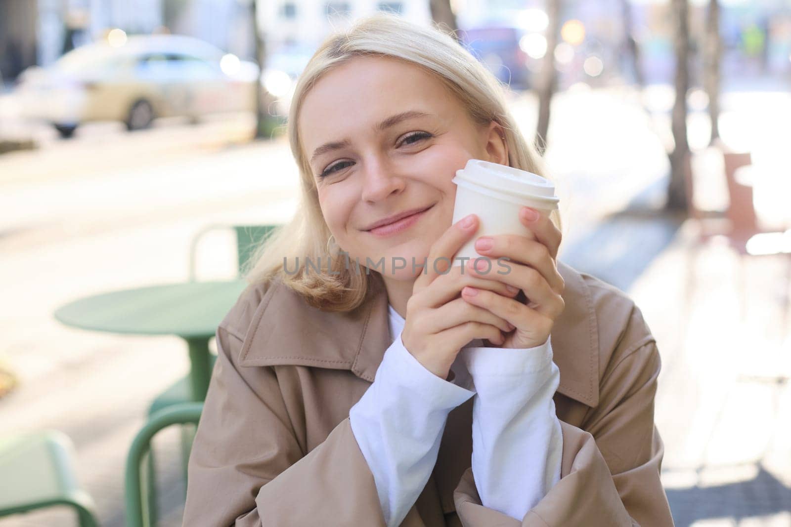 Close up portrait of smiling blond woman, holding cup of coffee, sitting in outdoor, street cafe, looking happy, waiting for someone in restaurant outside.