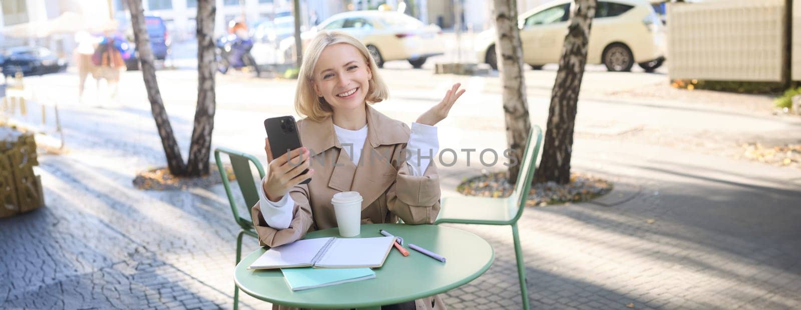 Happy young blond female model in coffee shop, talks on video chat, using smartphone and having conversation while resting outdoors in city centre.