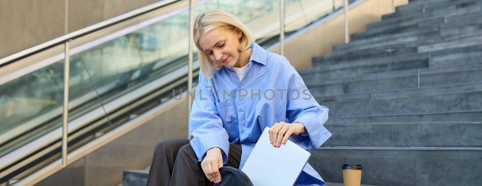 Smiling young female model, packing her laptop in backpack, sitting on city stairs outdoors, drinking cup of coffee before lecture in university.
