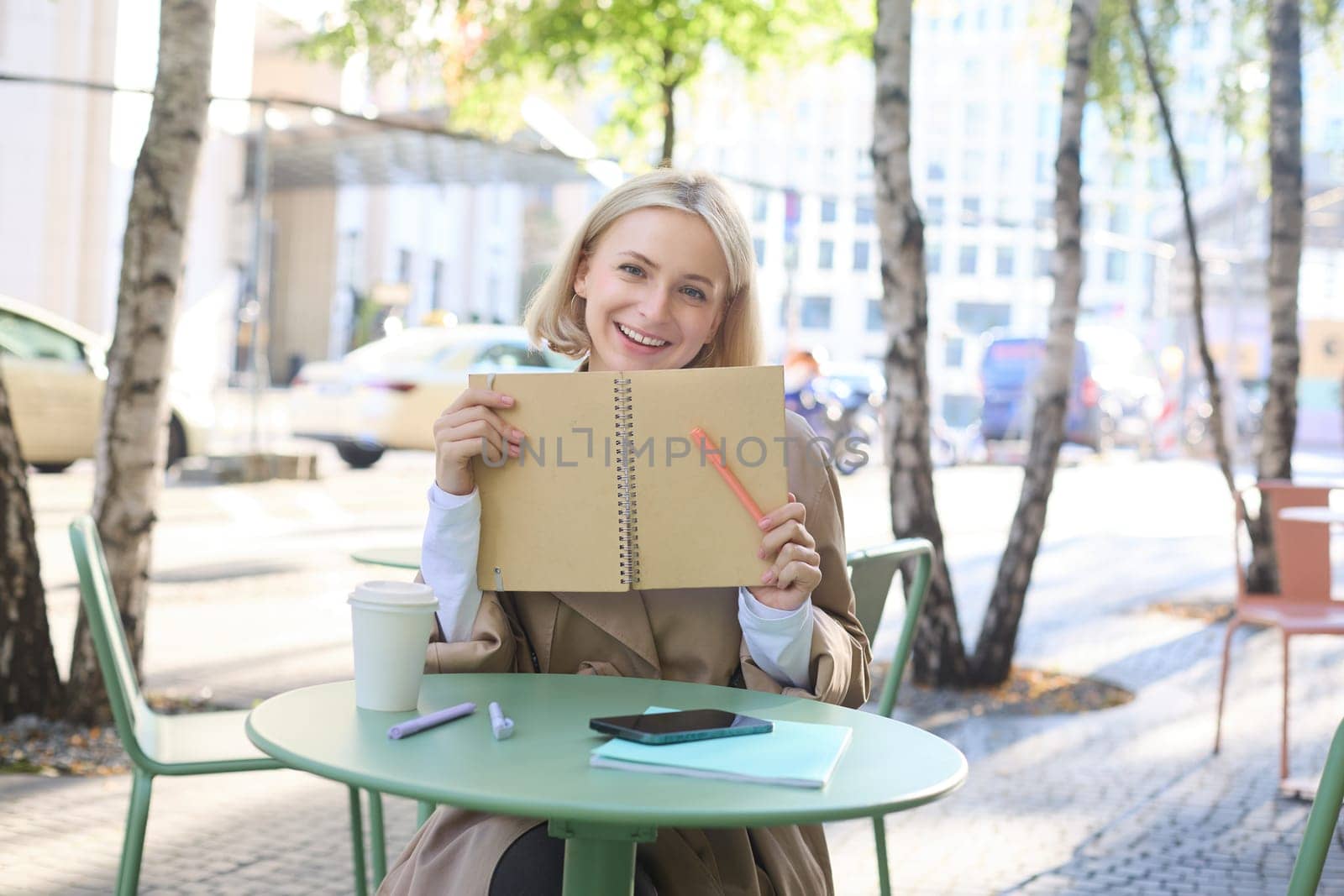 Portrait of carefree, smiling young woman with notebook and pen, sitting in outdoor cafe near table, writing in journal, making plans in her planner.