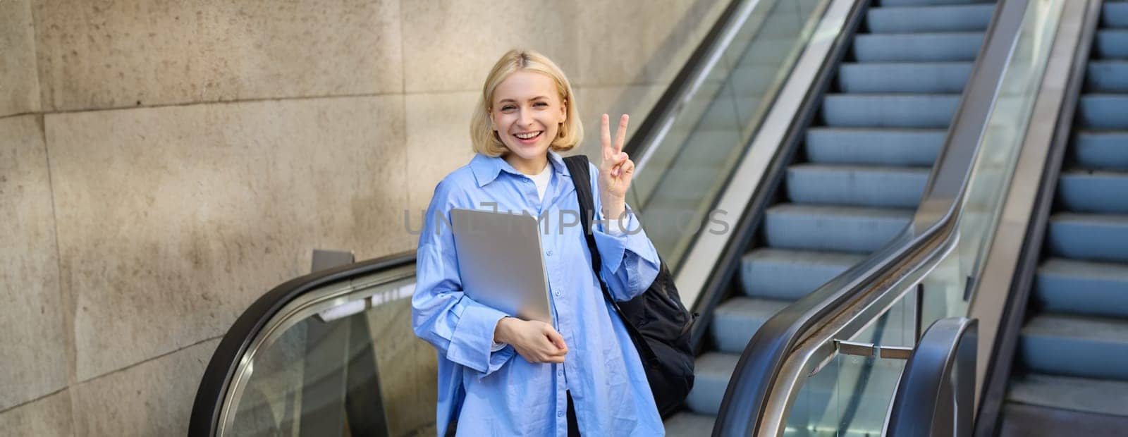 Beautiful young blond woman in blue collar shirt, going to work, using escalator, standing with laptop and showing peace sign by Benzoix