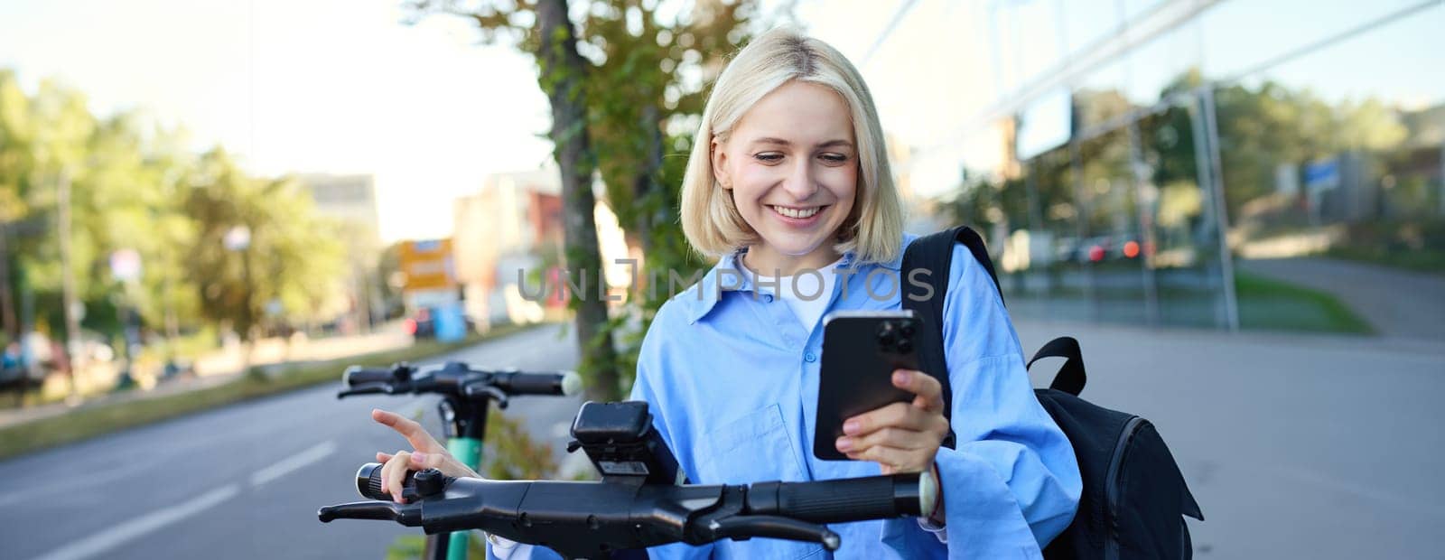 Portrait of young smiling female model, renting an electric scooter, using mobile phone to scan QR code with smartphone app, riding home from university, standing outdoors on street by Benzoix