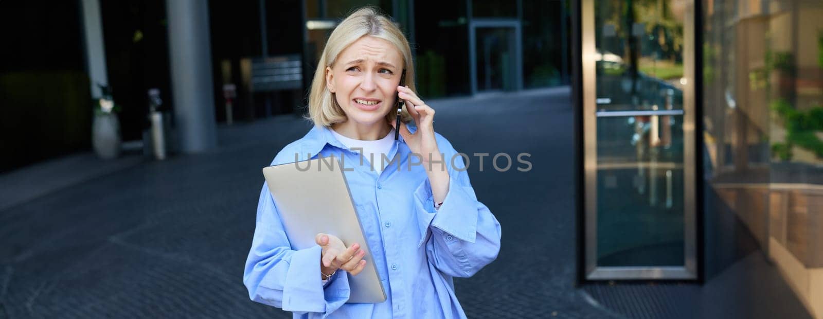 Image of young smiling woman, working in company, standing near office with laptop, answers phone call, chatting with someone.
