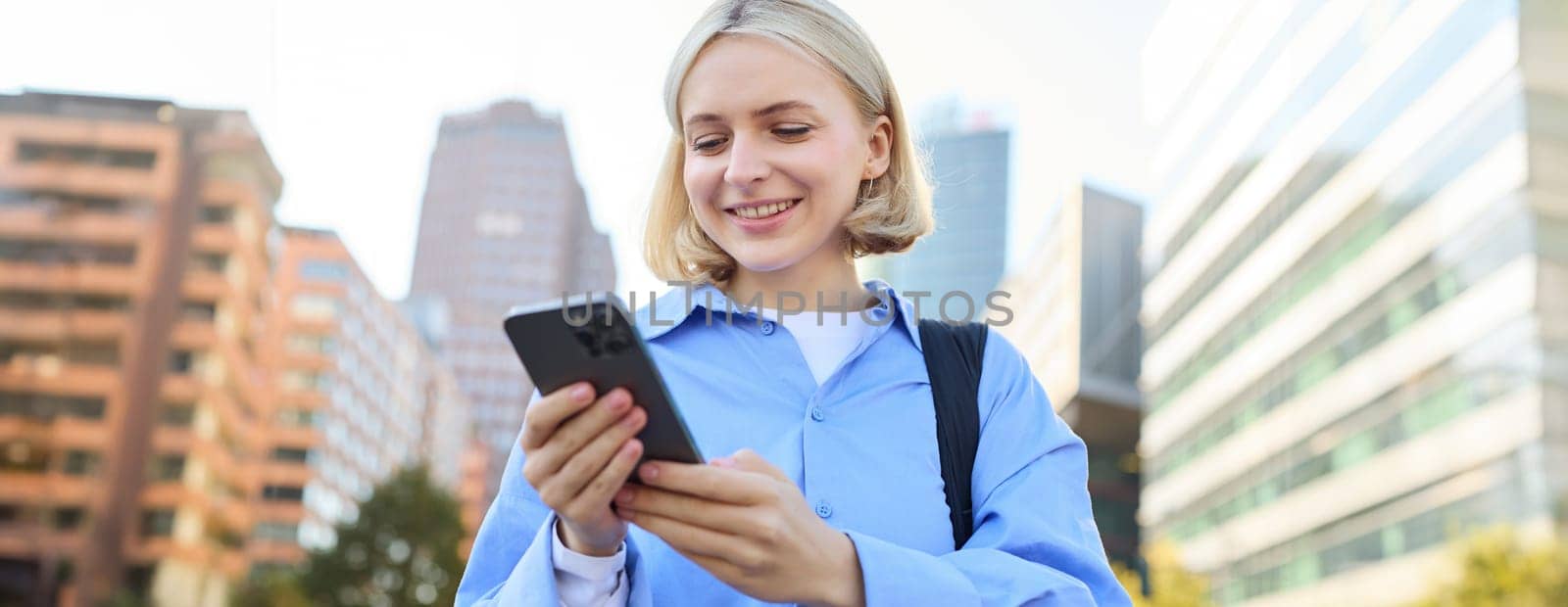 Close up portrait of stylish young blonde woman, standing on street, checking her mobile phone, using smartphone app to get around town, looking at online map for directions.