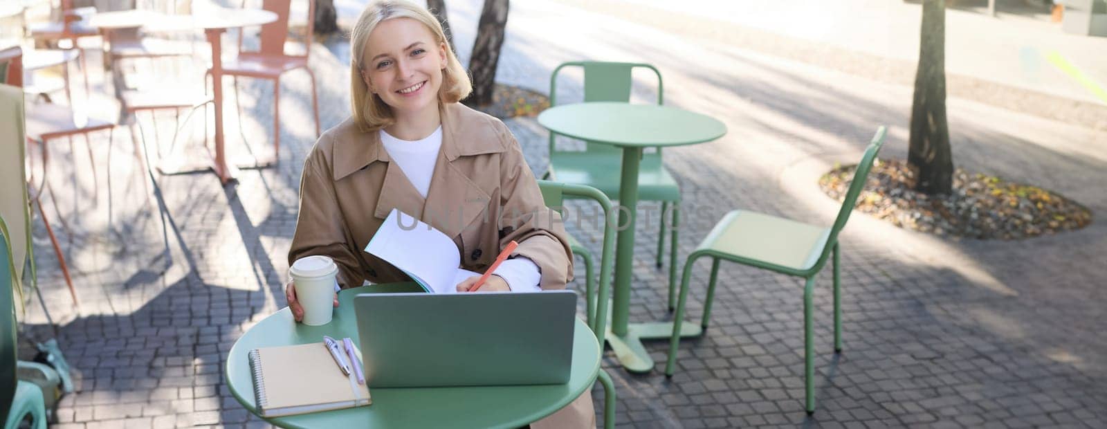 Portrait of young smiling blond woman, working on laptop, sitting in outdoor cafe on street, drinking coffee. Lifestyle and people concept