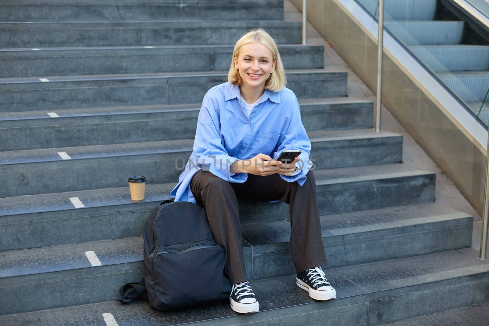 Portrait of woman on stairs, sitting with backpack, using mobile phone, messaging, scrolling social media on lunch break in university.