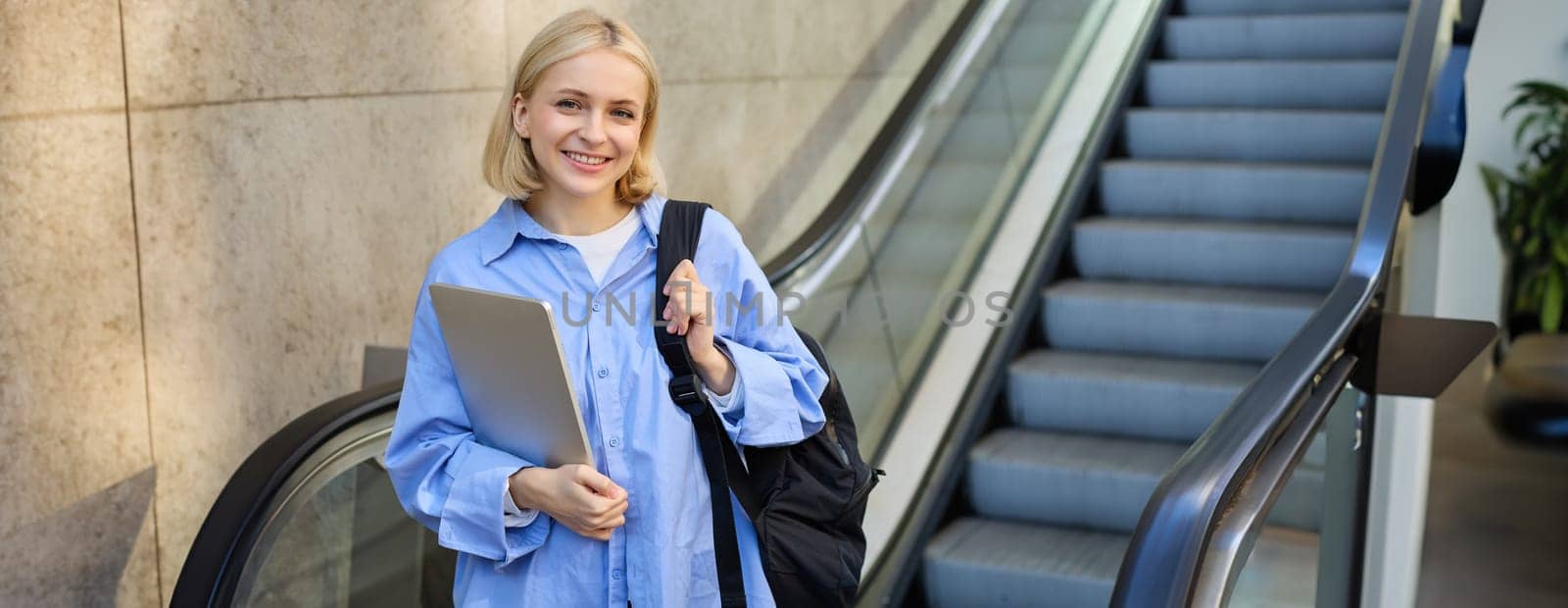 Concept of education and people. Young woman with backpack, carries laptop in hand, standing near escalator, going to tube, on her way to work on university.
