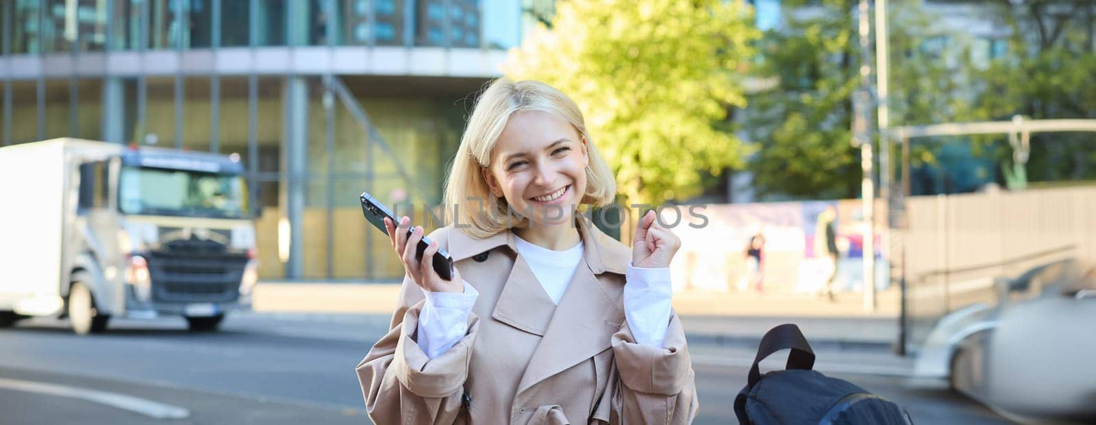 Cheerful blonde woman, sitting on bench and celebrating victory, holding mobile phone, smiling pleased, triumphing with joy, receive good news.