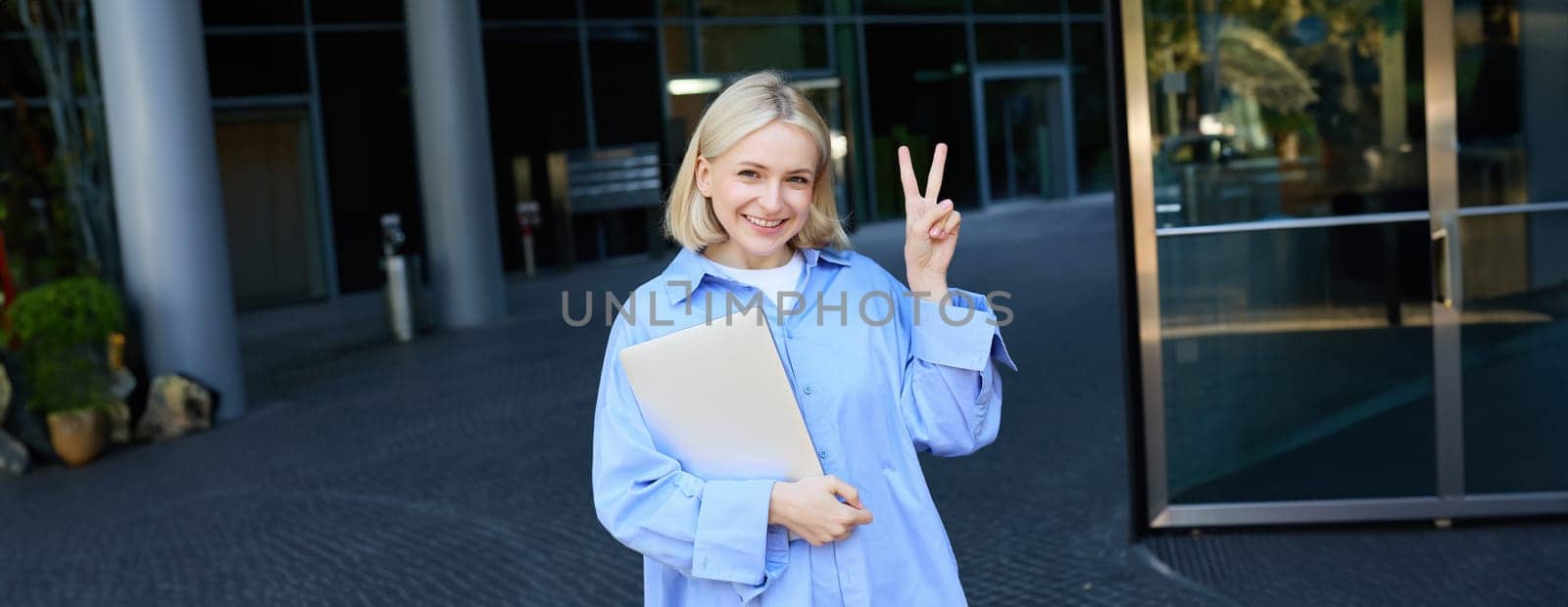 Young positive female model, student with notebooks and study material showing peace sign, standing near university, college campus, promo for new education scheme.