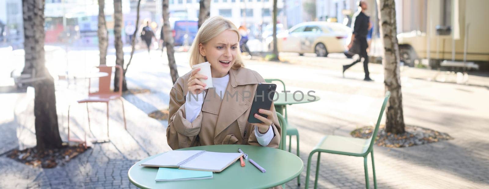 Portrait of cheerful, beautiful young woman taking selfie, chatting on video using smartphone, posing outdoors in coffee shop.