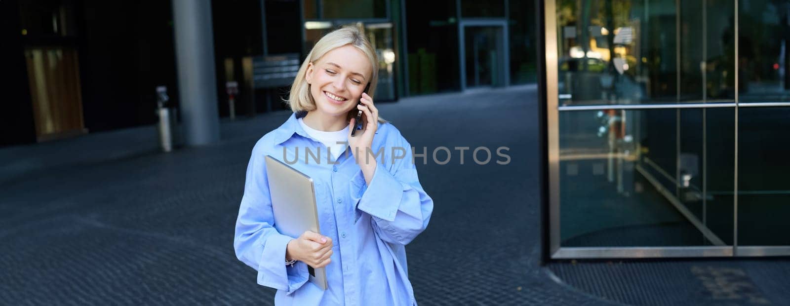 Portrait of young businesswoman in blue collar shirt, talking on mobile phone, standing near office building and smiling, holding work laptop.