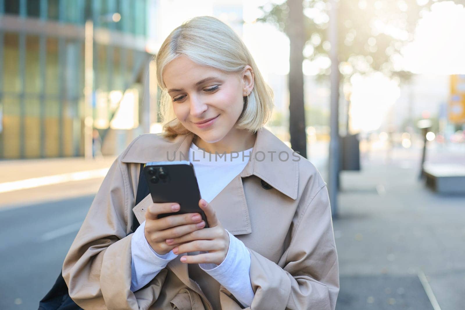 Image of young modern woman, student walking on street and looking at her mobile phone, smiling and looking happy. Copy space