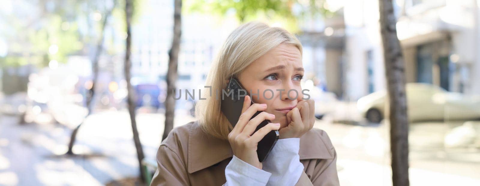 Image of young stressed, upset woman, talking on mobile phone with worried face expression, has difficult conversation over the telephone, sitting in outdoor cafe.