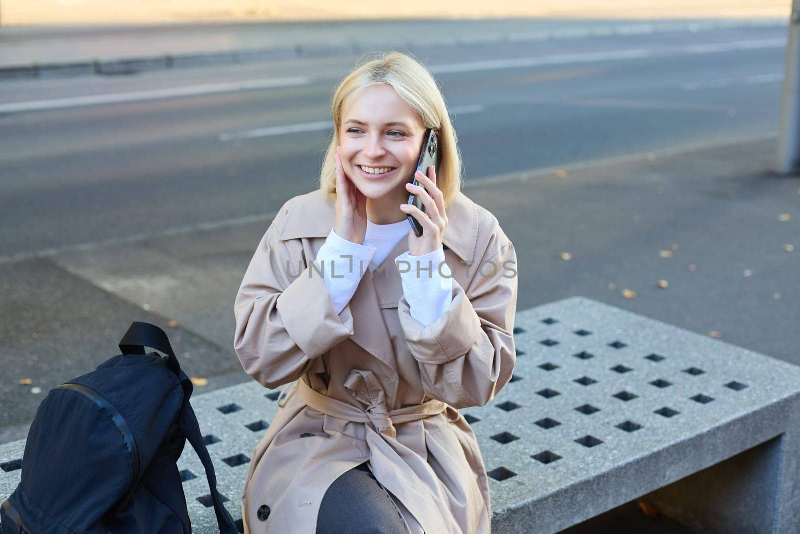 Image of young blond woman sitting on street bench with backpack, talking on mobile phone, answers a call and smiling while chatting with someone.