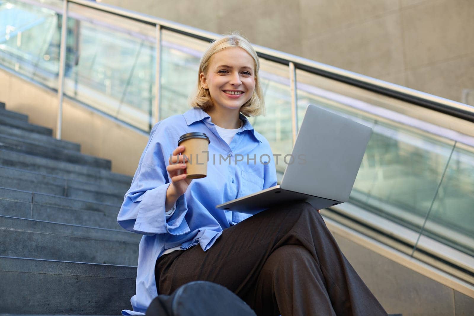 Image of stylish young modern woman, student doing homework, studying outdoors on campus stairs, sitting with laptop and coffee, drinking her cappuccino and connecting to public wifi by Benzoix