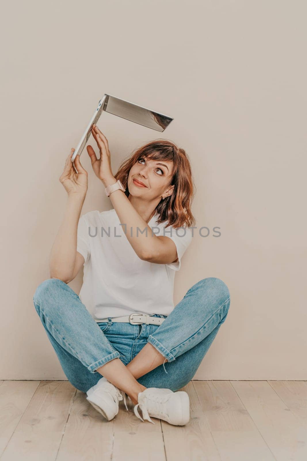 A brunette sits on the floor with a laptop on a beige wall background. She is wearing a white T-shirt, jeans and white sneakers