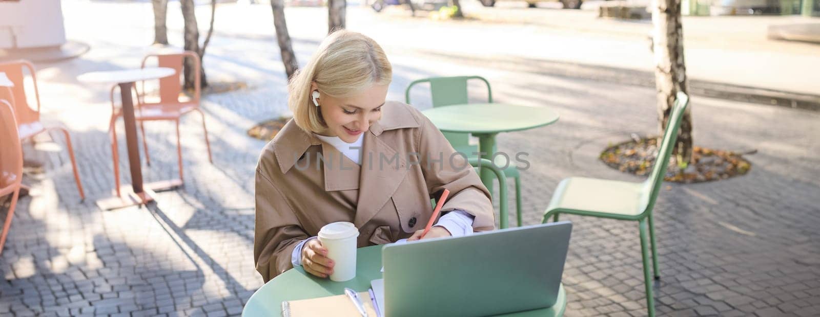 Portrait of young blond woman, female student in street cafe, wearing wireless headphones, using laptop, having online meeting, attend web lecture or course, doing homework by Benzoix