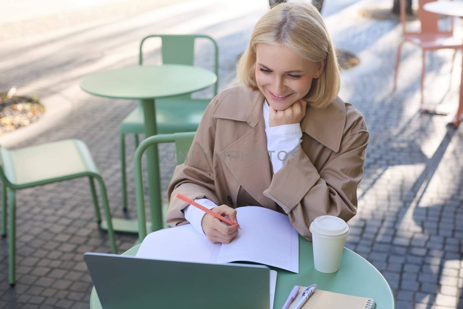 Image of young woman studying outdoors, sitting in cafe, making notes while listening to online lecture using laptop, making notes in notebook, writing.