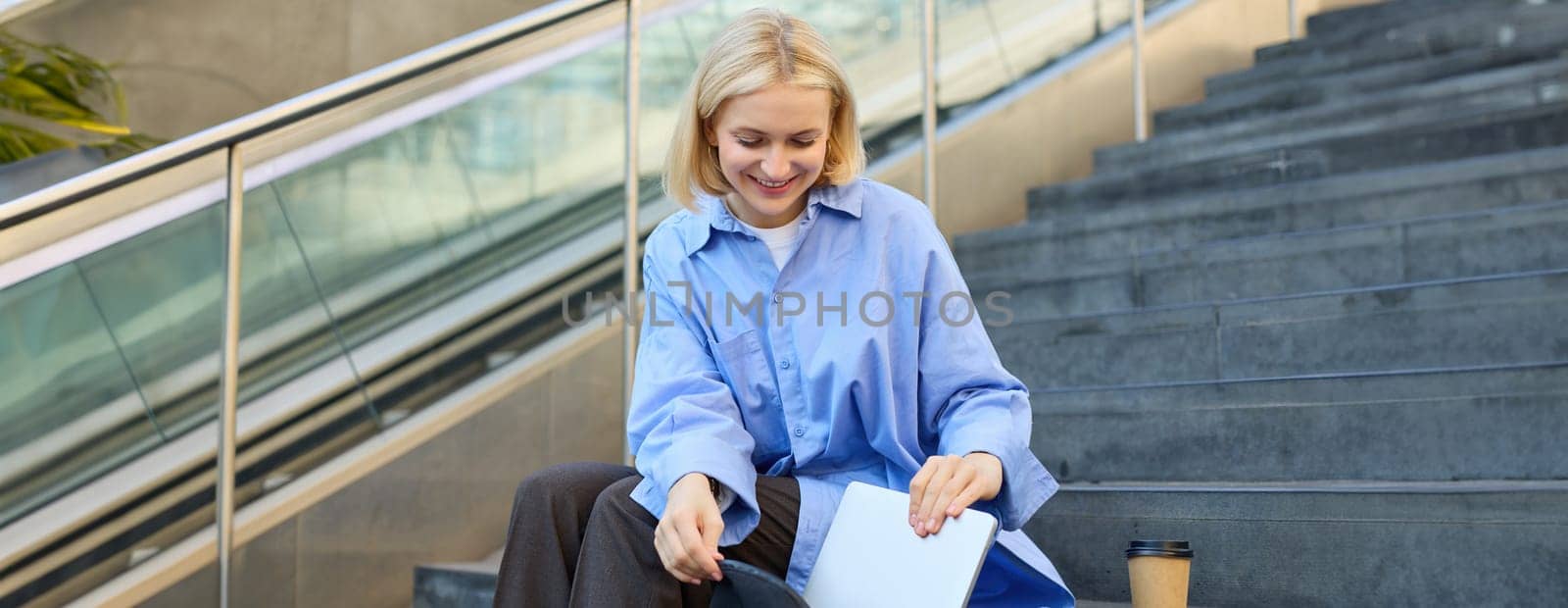 Image of stylish young woman, putting away her laptop in backpack, sitting on stairs near campus, packing for university, drinking coffee by Benzoix