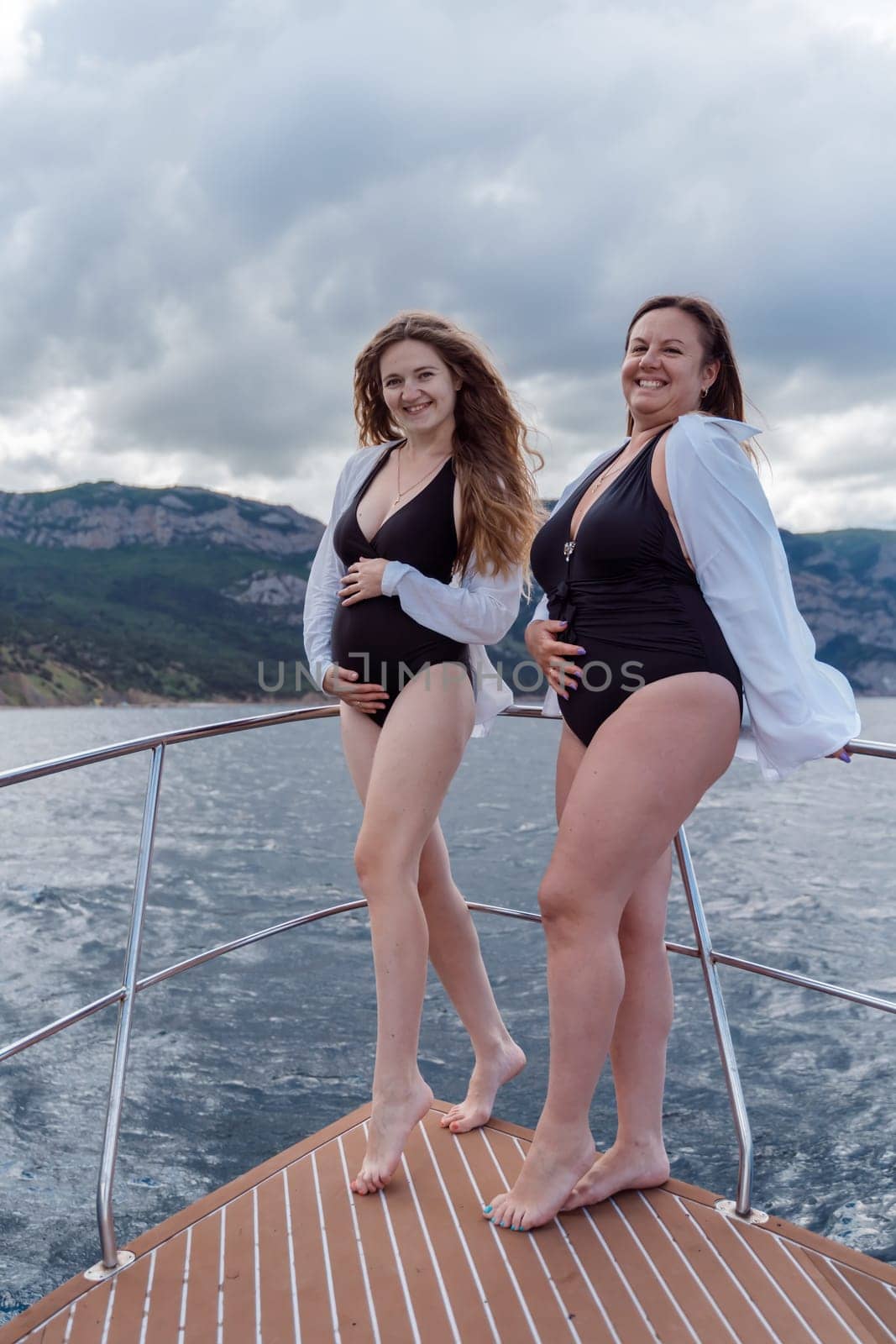 Pregnant on a yacht. Happy models in a swimsuit posing on a yacht against sky with clouds and mountains