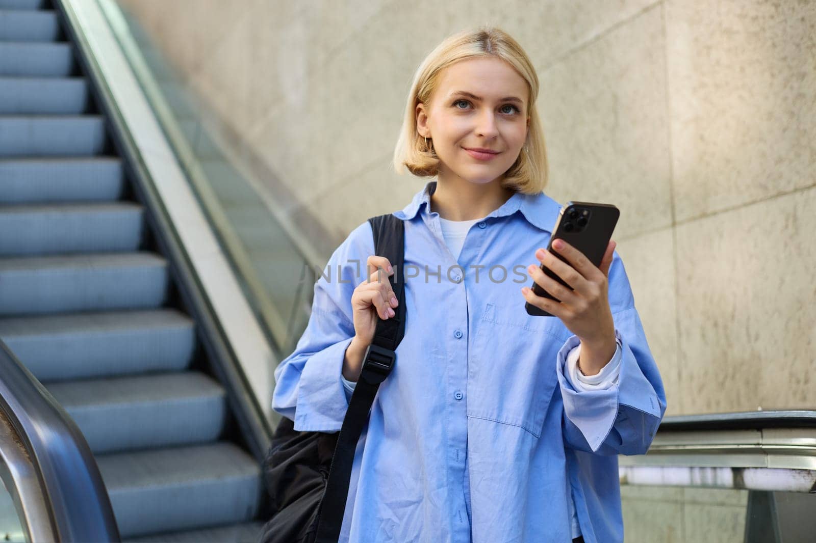 Portrait of young college student, woman with backpack and mobile phone, standing near escalator, reading notification on smartphone.