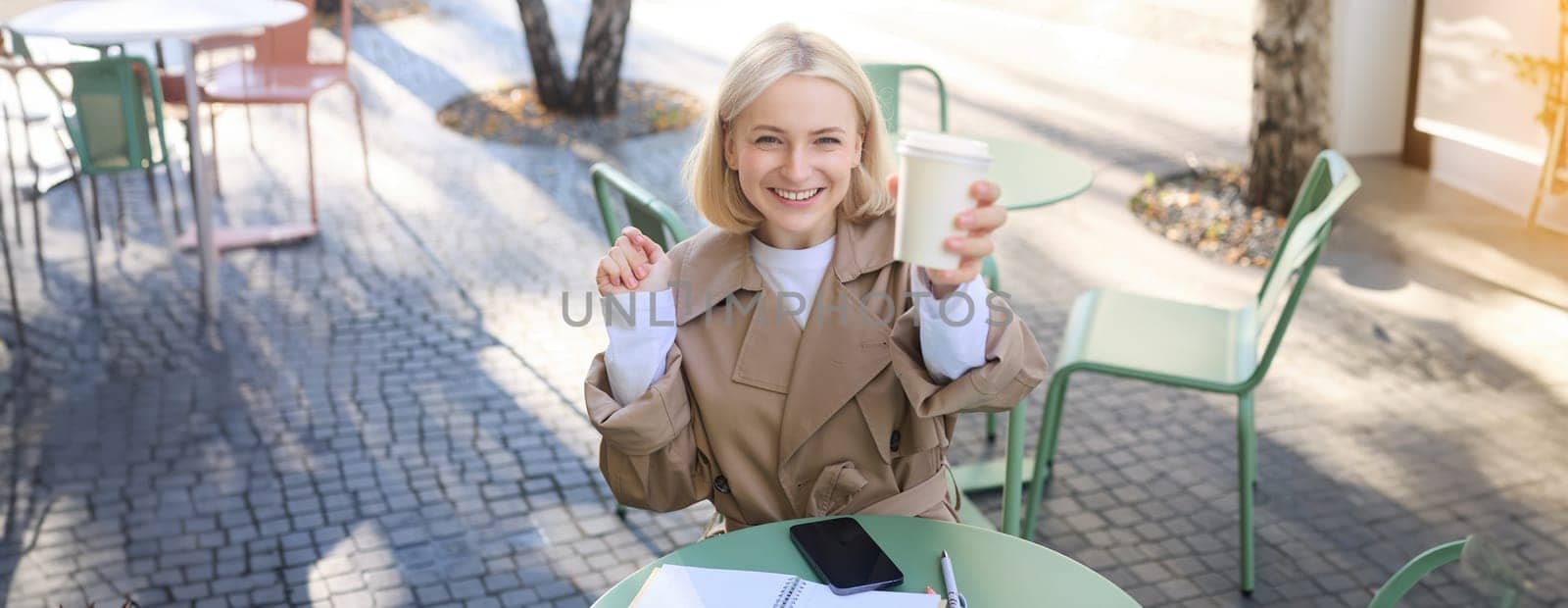 Beautiful blond woman, smiling, showing white takeaway coffee cup, drinking beverage in cafe shop, sitting outdoors.