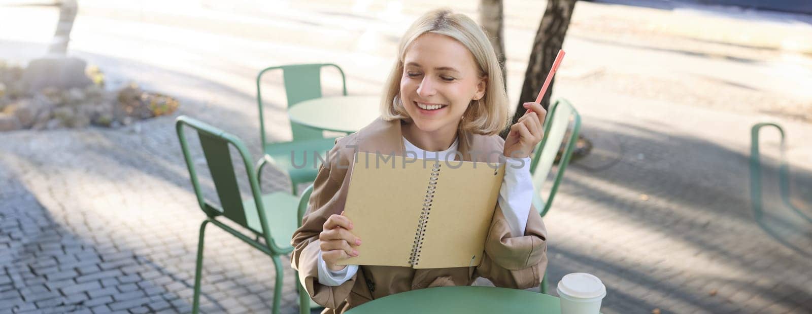 Portrait of laughing, smiling young woman, modern creative girl with notebook and pen, sitting in outdoor coffee shop and drawing, writing in journal, working on project.