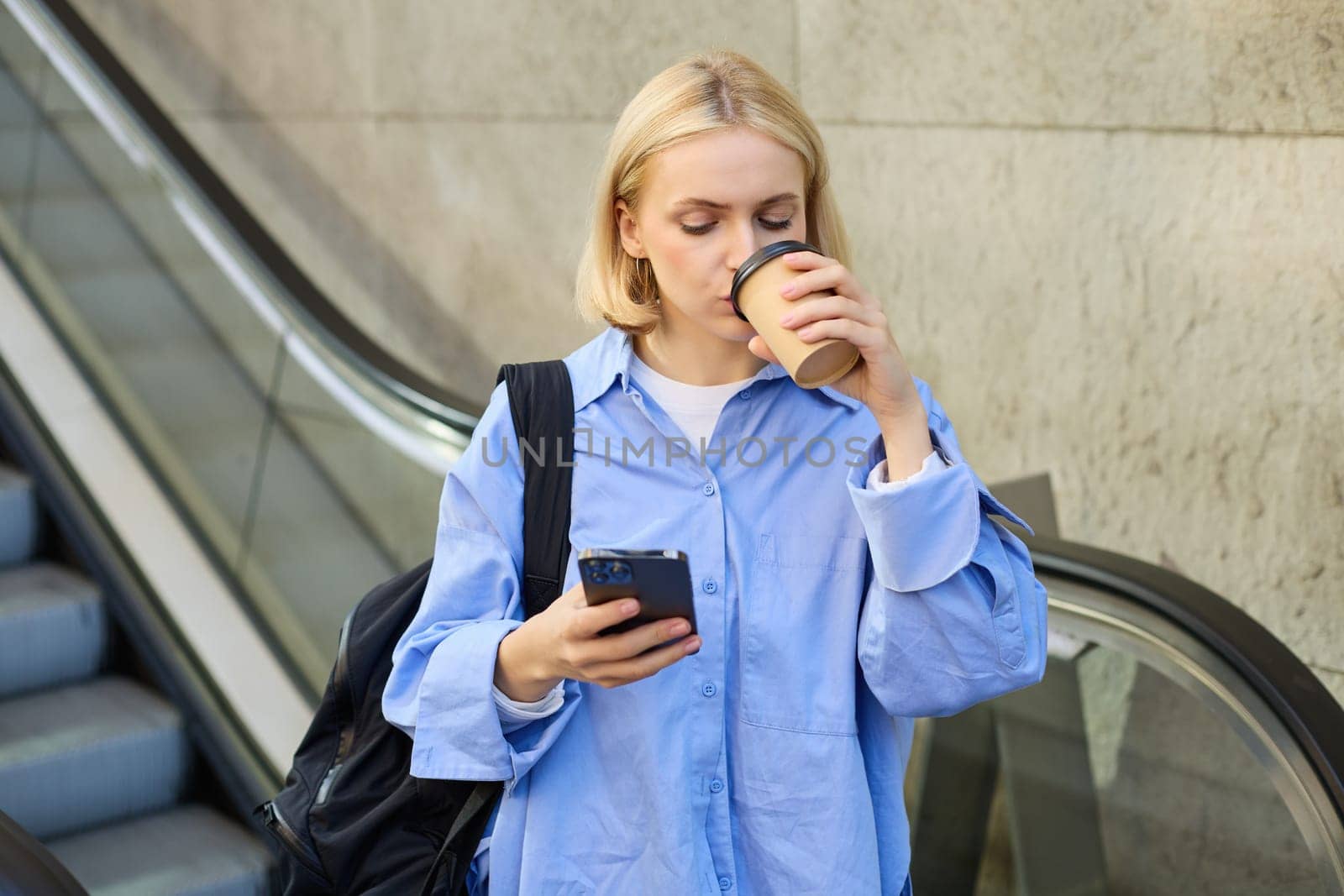 A happy, elegant businesswoman descending the escalator and holding takeaway coffee while reading important messages. A woman on the escalator with a phone.