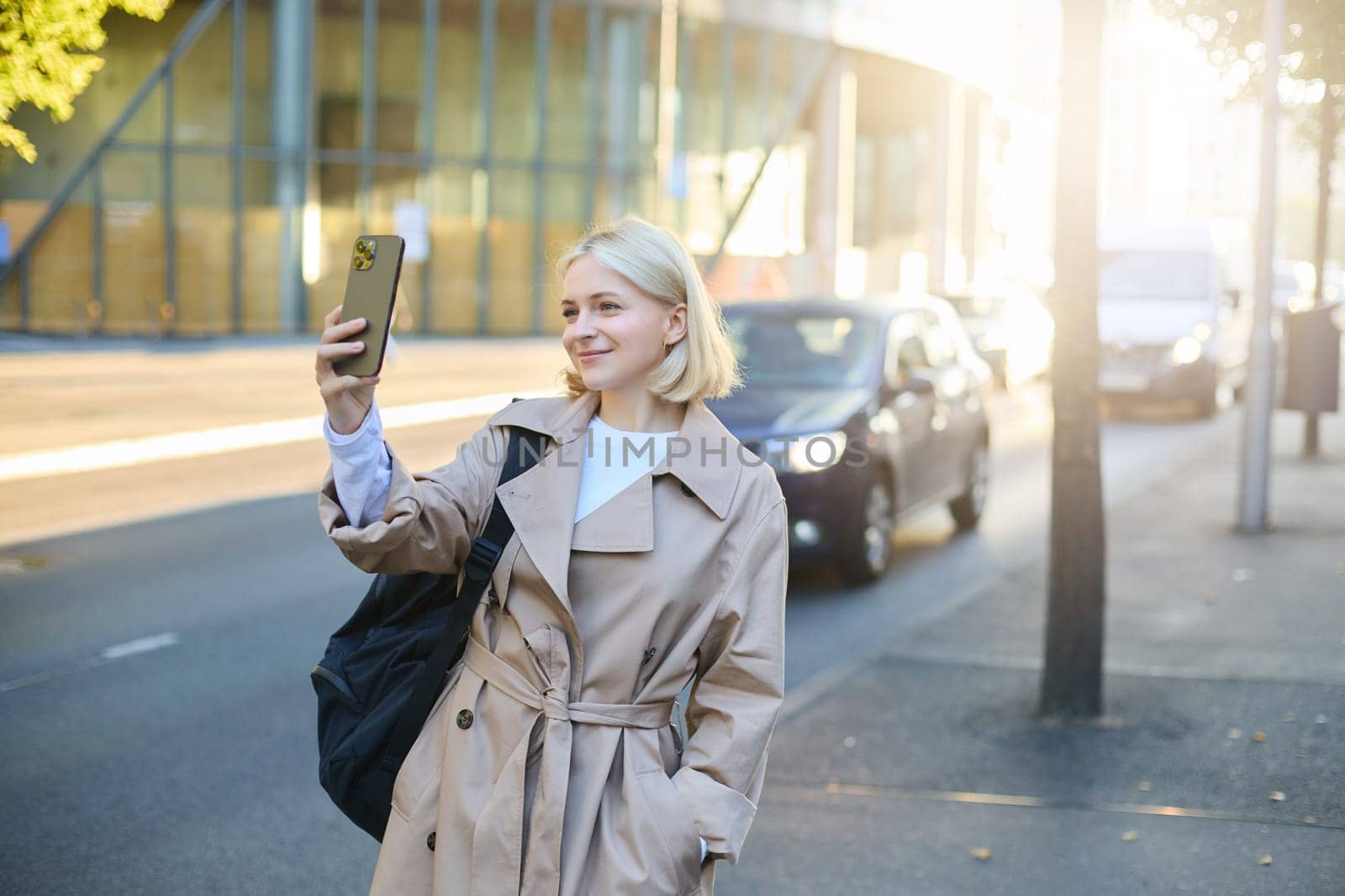 Image of stylish smiling woman, blogger taking selfie on street, smiling and posing for social media photo on smartphone, using mobile phone camera.