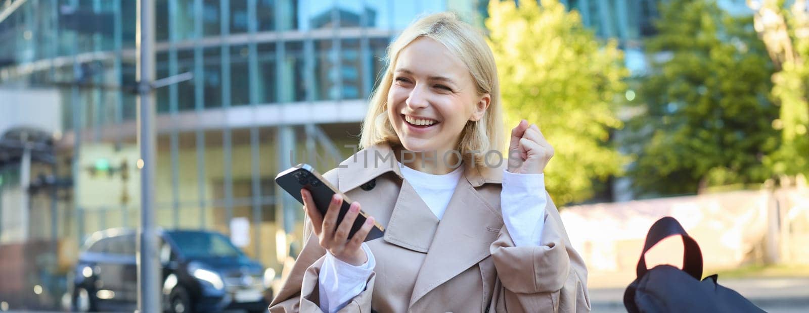 Upbeat young woman celebrating, winning and triumphing, sitting on street bench and laughing, holding mobile phone by Benzoix