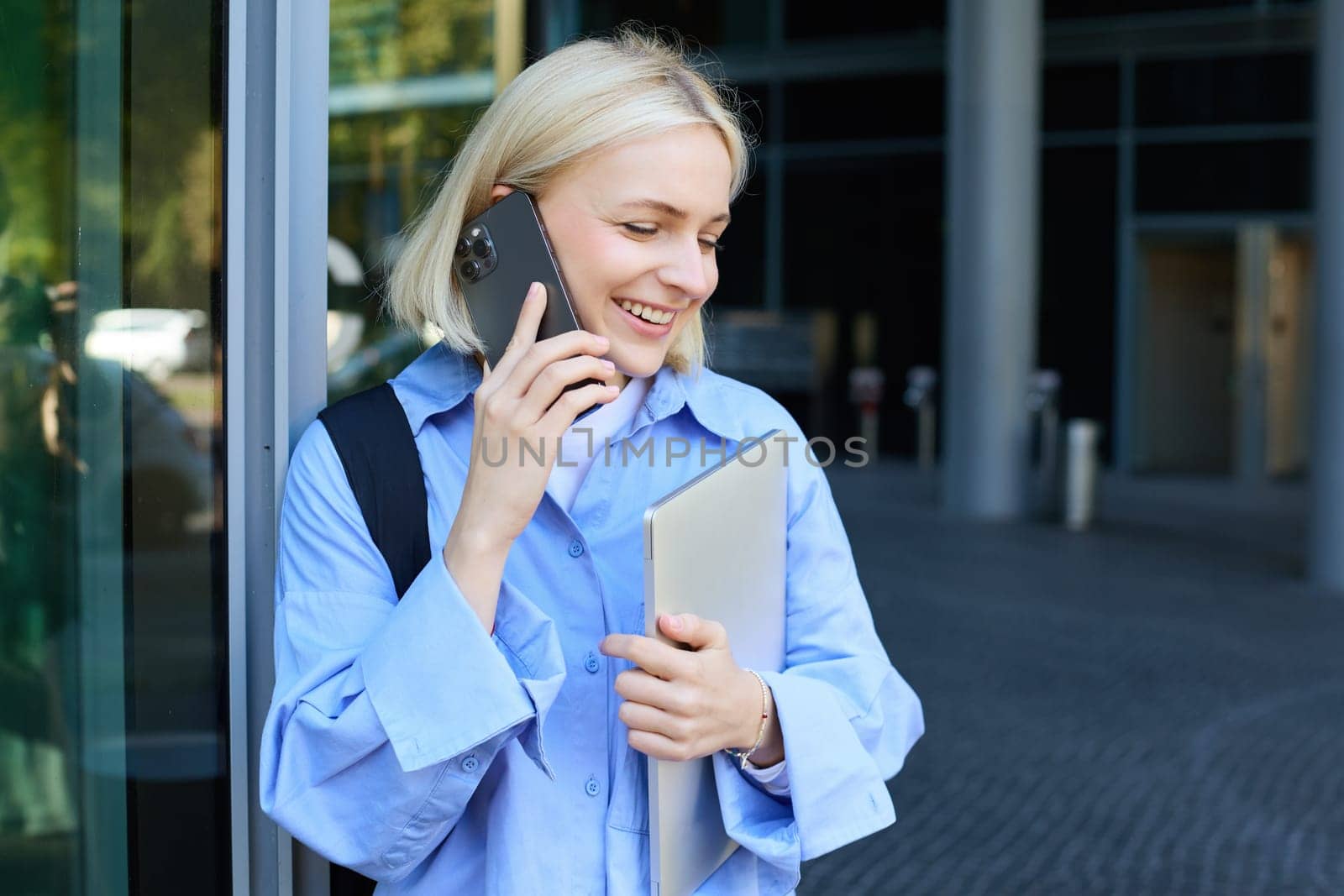 Young modern woman, female employee, standing near office building, answers phone call, talking, holding laptop and backpack, smiling while having a nice, friendly chat.