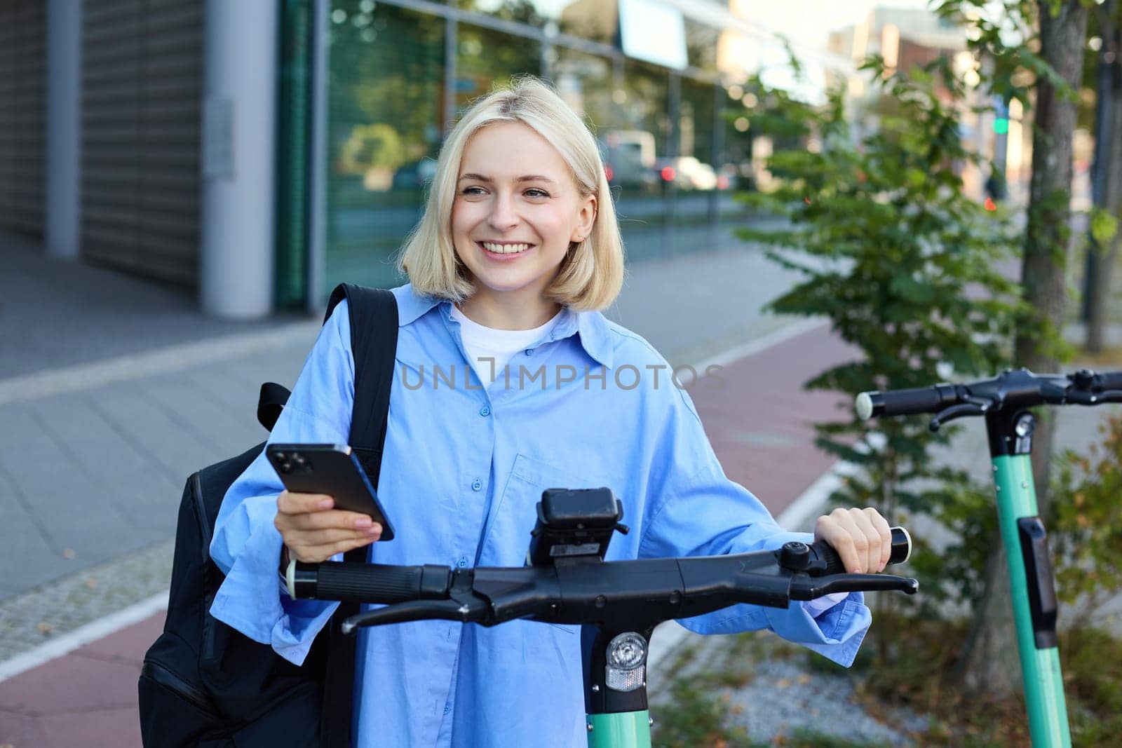 Portrait of young smiling woman, unlocks electric scooter, scans qr code with smartphone app to unlock her ride, standing on street near building.