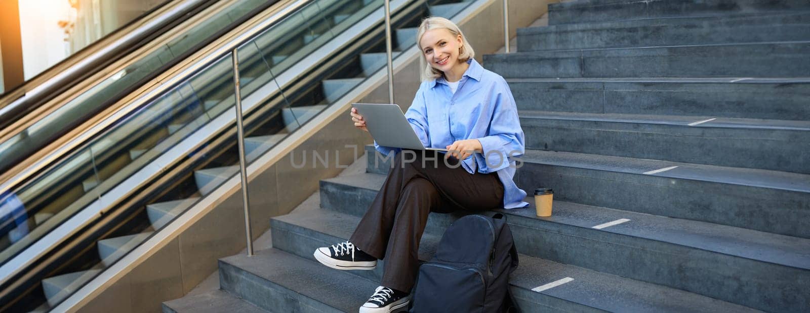 Image of young woman, student studying online, working remotely, sitting on stairs with laptop and drinking coffee in takeaway cup, elearning by Benzoix