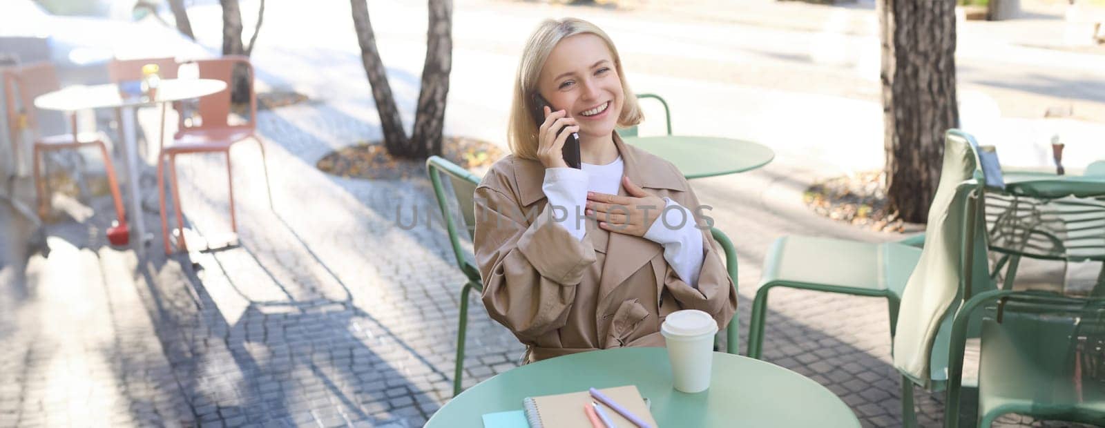 Image of stylish blond woman talking on mobile phone, drinking coffee in cafe outdoors, enjoying warm weather in city centre, answer telephone call on smartphone.