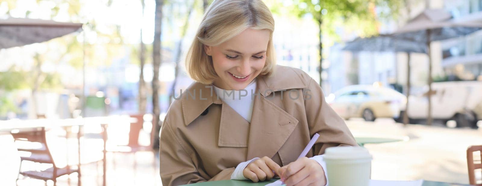 Image of young modern woman, student working on project, sitting in outdoor cafe, drinking coffee and working alone, studying or doing homework.