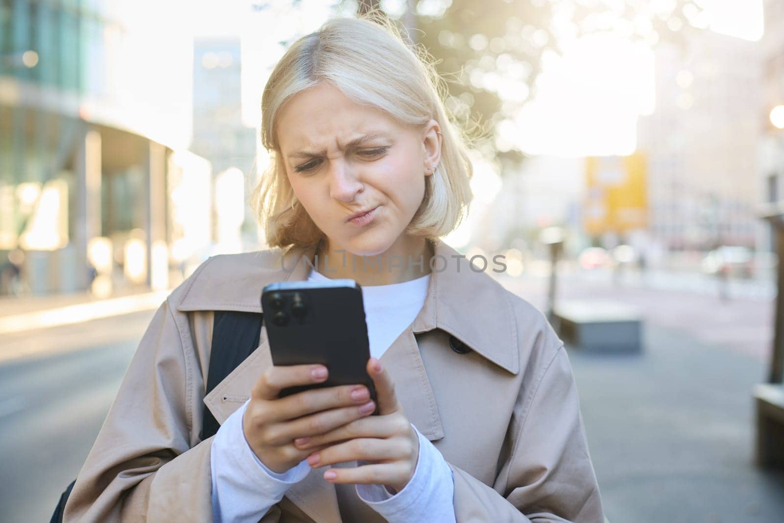 Close up portrait of woman looking sceptical at her mobile phone, standing on street, frowning and standing perplexed, reading something on smartphone screen by Benzoix