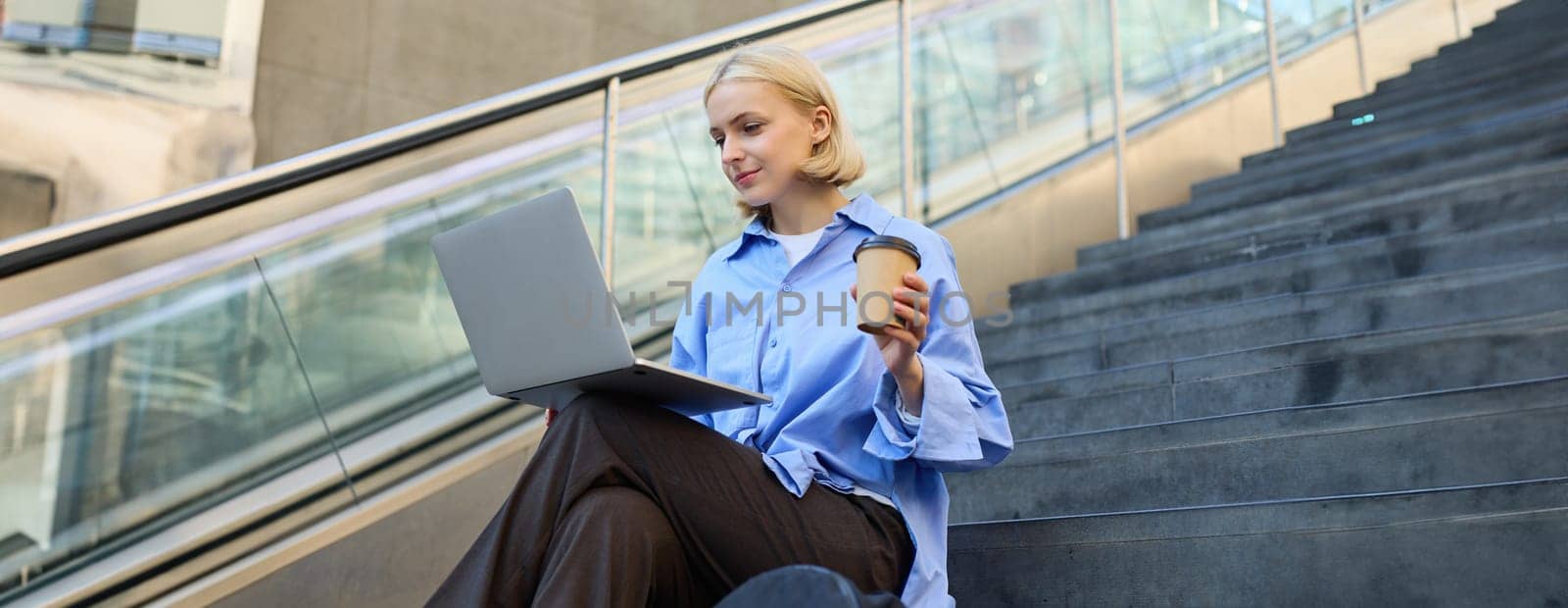 Image of stylish young modern woman, student doing homework, studying outdoors on campus stairs, sitting with laptop and coffee, drinking her cappuccino and connecting to public wifi by Benzoix