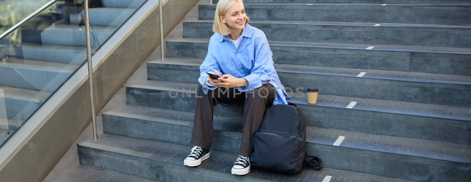 Portrait of woman on stairs, sitting with backpack, using mobile phone, messaging, scrolling social media on lunch break in university by Benzoix