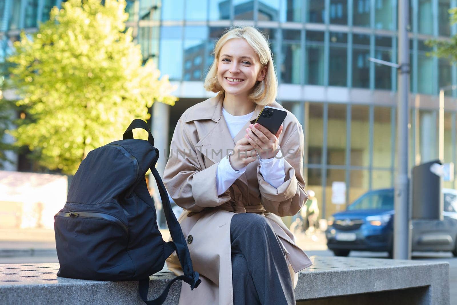 Outdoor shot of young blonde woman on street, sitting on bench with mobile phone, smiling at camera, waiting for friend outside of building, having a break.