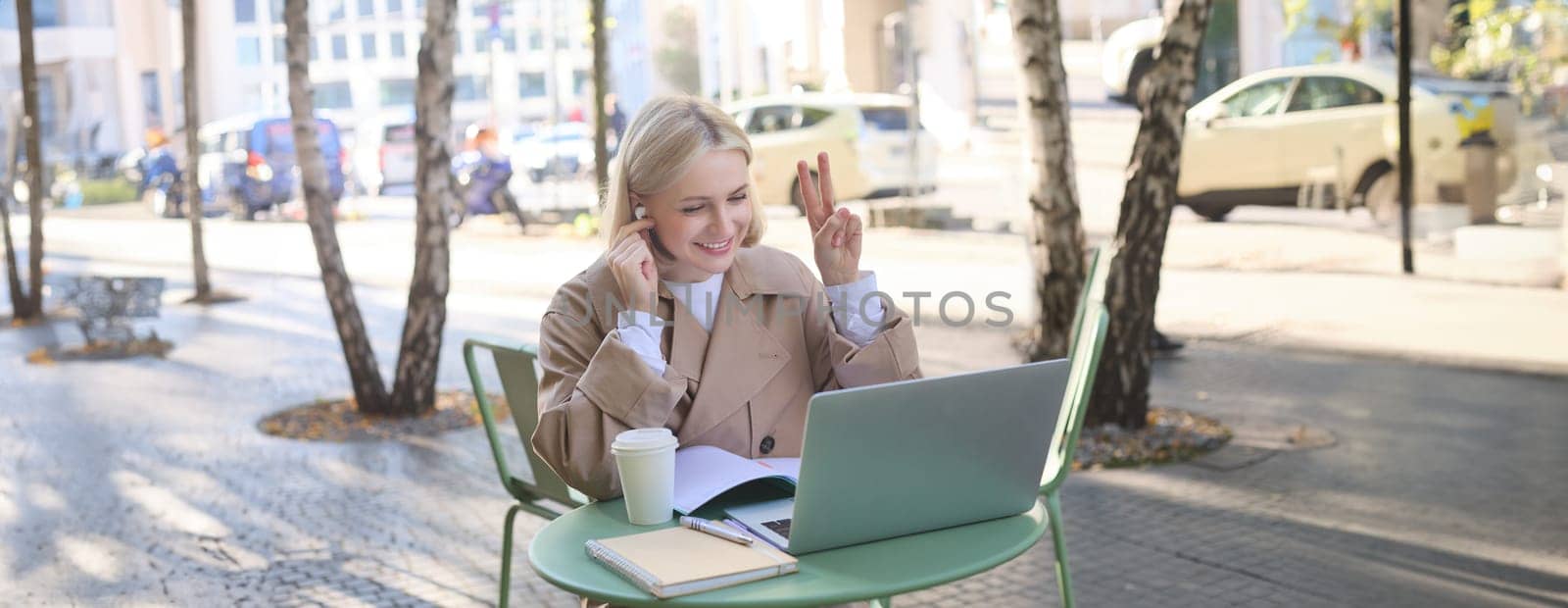 Image of smiling, beautiful young woman in wireless headphones, connects to online meeting, video chatting in outdoor coffee shop, showing peace sign, using laptop, studying remotely by Benzoix
