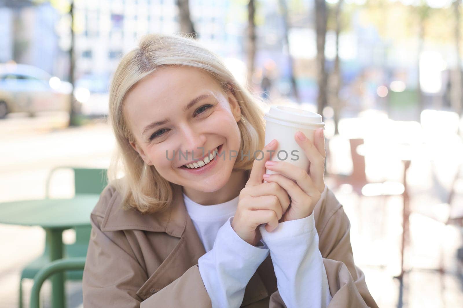 Close up portrait of smiling blonde woman, university student, holding cup of coffee, warming up her hands on chilly autumn day, sitting in outdoor cafe, laughing at camera.