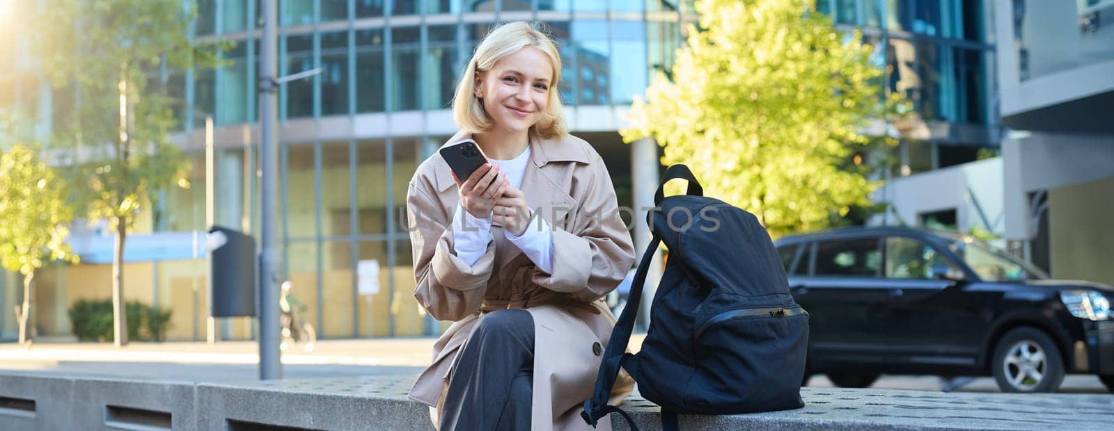 Lifestyle portrait of cute blonde girl with smartphone, sitting on bench with backpack, using mobile phone, reading message.