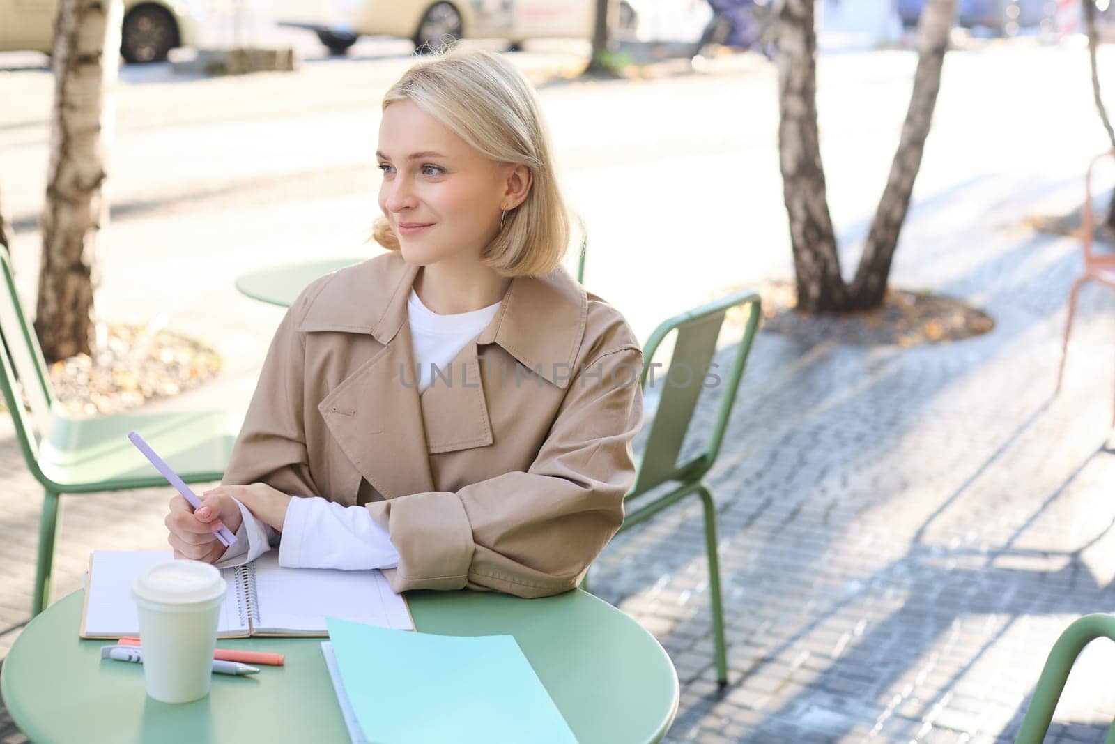 Image of young modern woman, student working on project, sitting in outdoor cafe, drinking coffee and working alone, studying or doing homework.