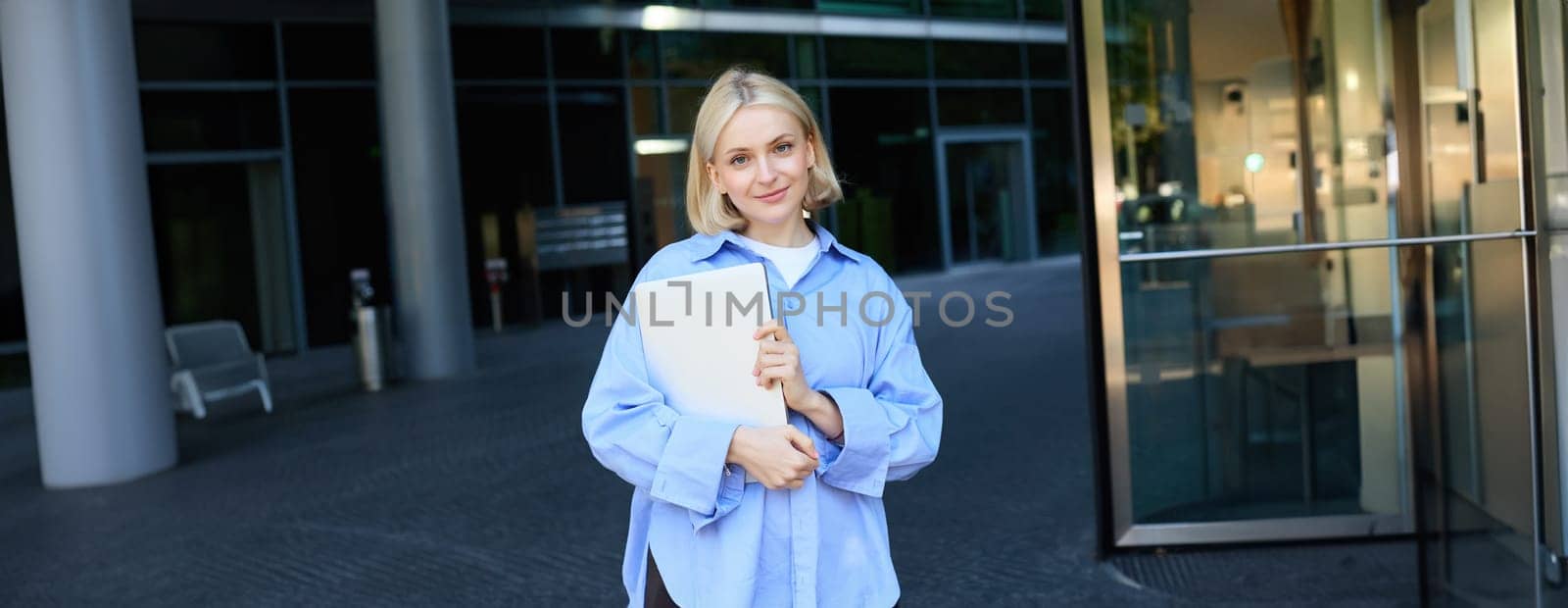 Image of young female employee, working woman standing near her office on street, holding pile of documents and smiling. People and lifestyle concept