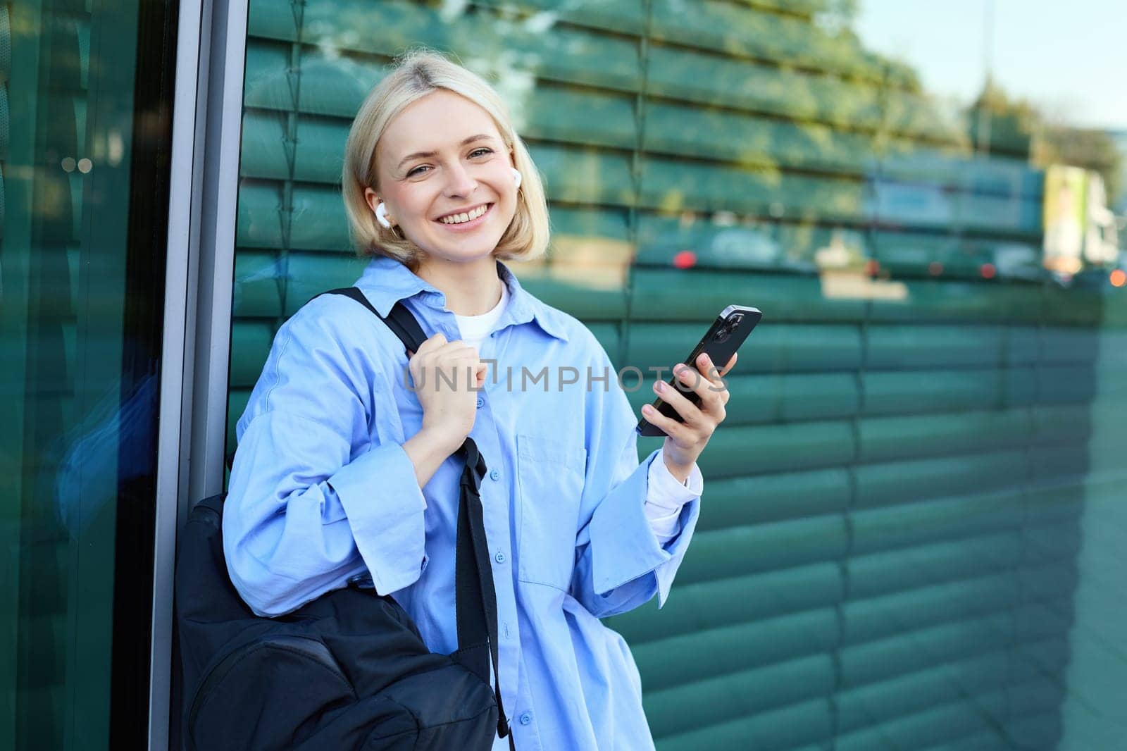 Lifestyle portrait of smiling young female model, student with backpack, waiting for someone on street, standing outdoors with smartphone by Benzoix