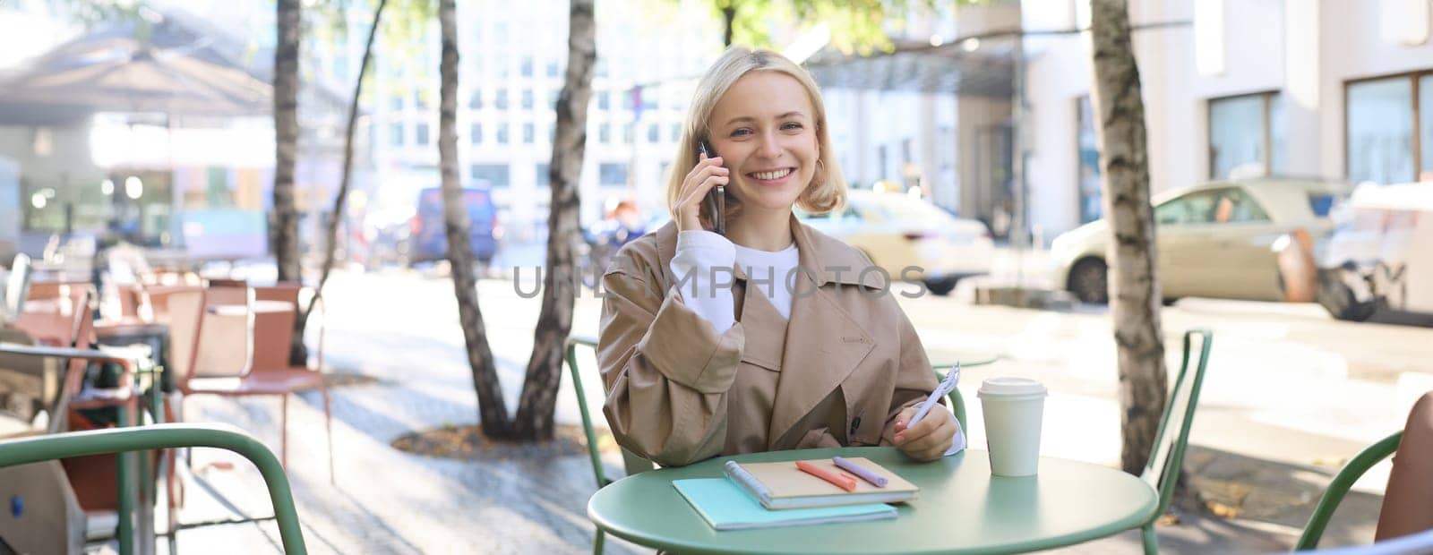 Friendly young woman in trench, sitting outdoors on sunny day, spending time in cafe, answer phone call, talking on mobile.