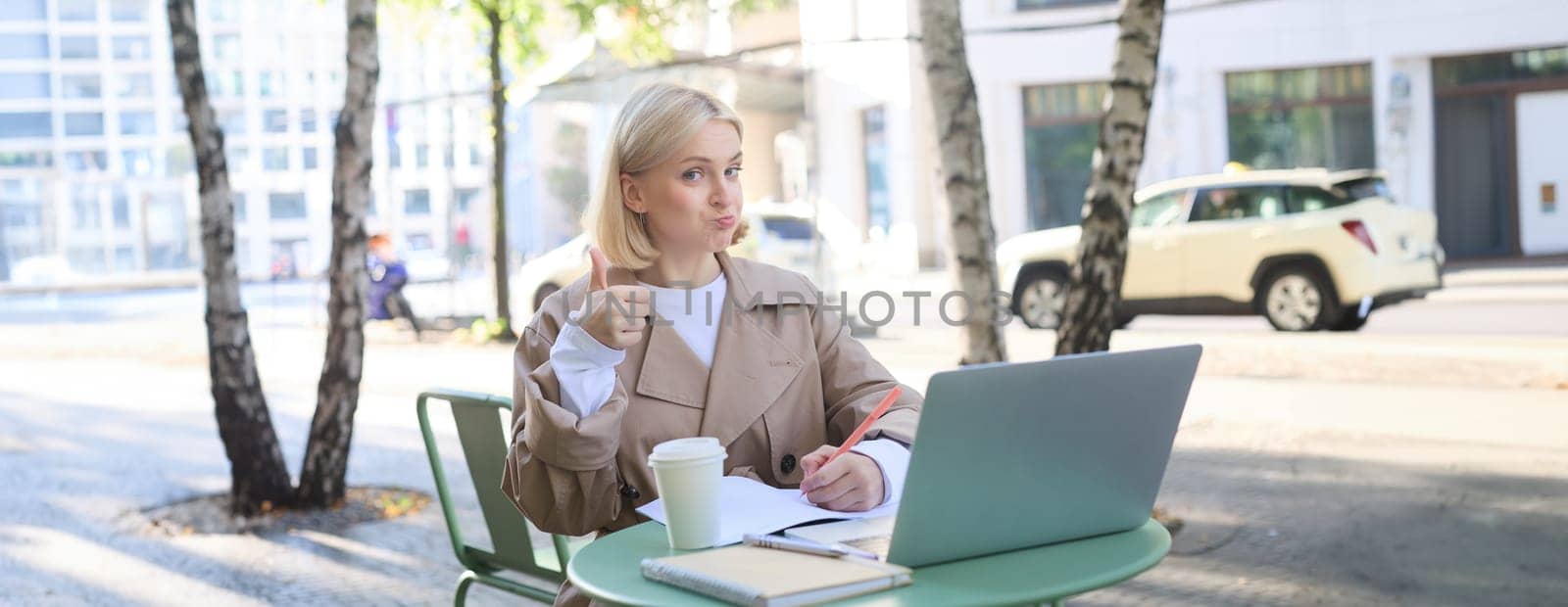 Image of impressed, satisfied young woman, sitting in outdoor cafe with laptop, showing thumbs up and nod in approval by Benzoix