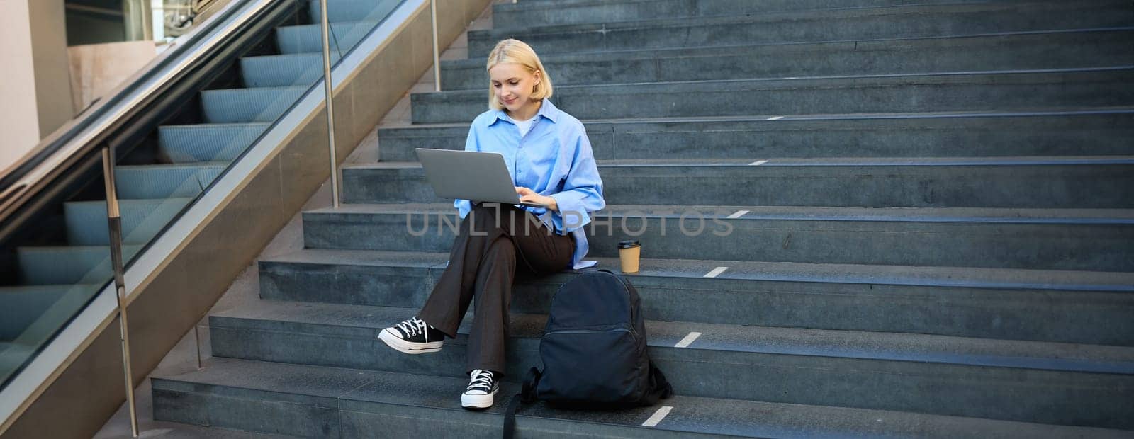 Modern young woman, student working on laptop, sitting outside campus in stairs with backpack and takeaway coffee, drinking her beverage and studying.